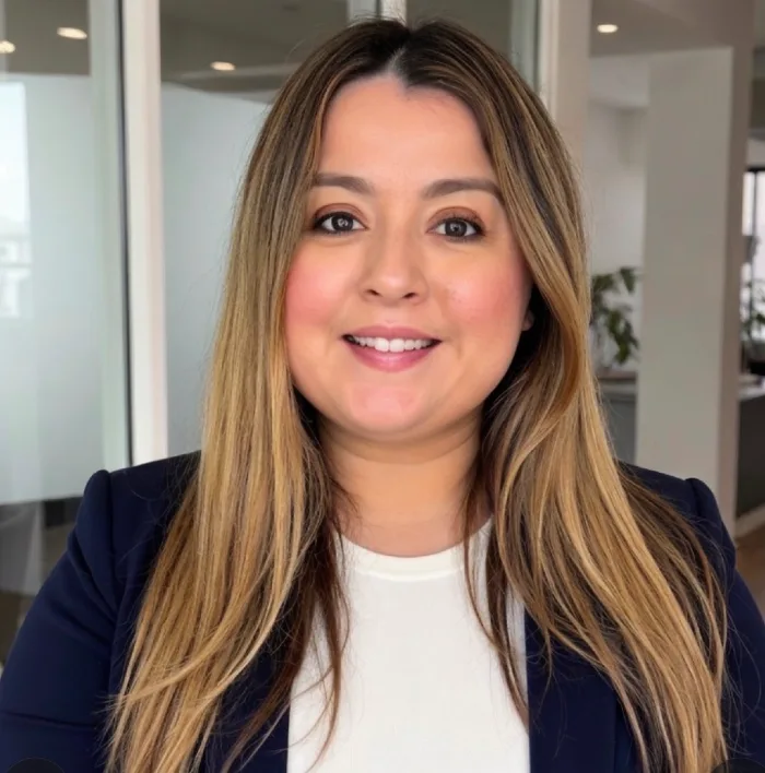 Professional headshot of a smiling woman with long light brown hair, wearing a navy blazer and white top in an office setting.