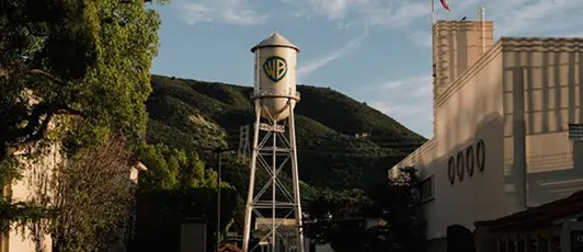 Water tower with Warner Bros. logo near buildings and green hills under a blue sky.