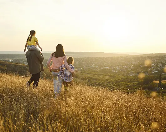 Family of four walking through a golden grassy field at sunset overlooking a distant town.