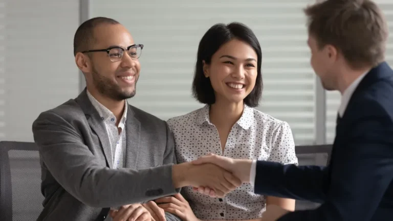 Three professionals, two men and one woman, smiling and shaking hands in office setting.