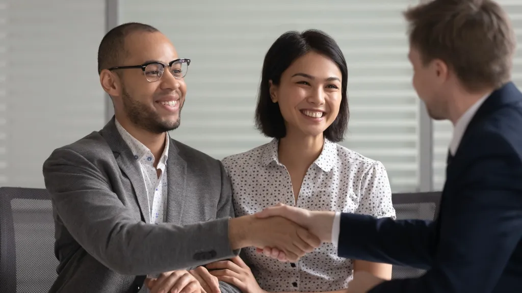 Three professionals, two men and one woman, smiling and shaking hands in office setting.