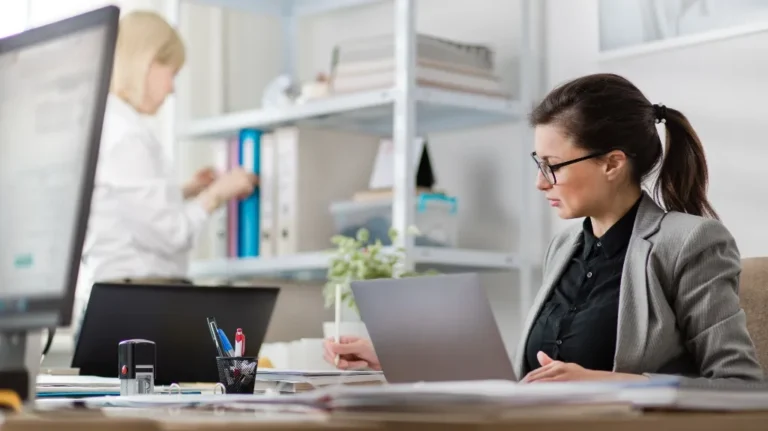 Two professional women working in a modern office with laptops and documents.