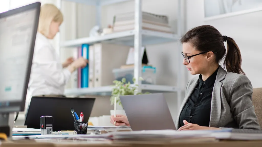 Two professional women working in a modern office with laptops and documents.