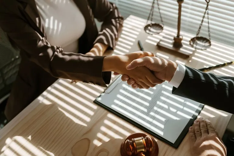 Two professionals shaking hands over a desk with legal scales, gavel, and documents.