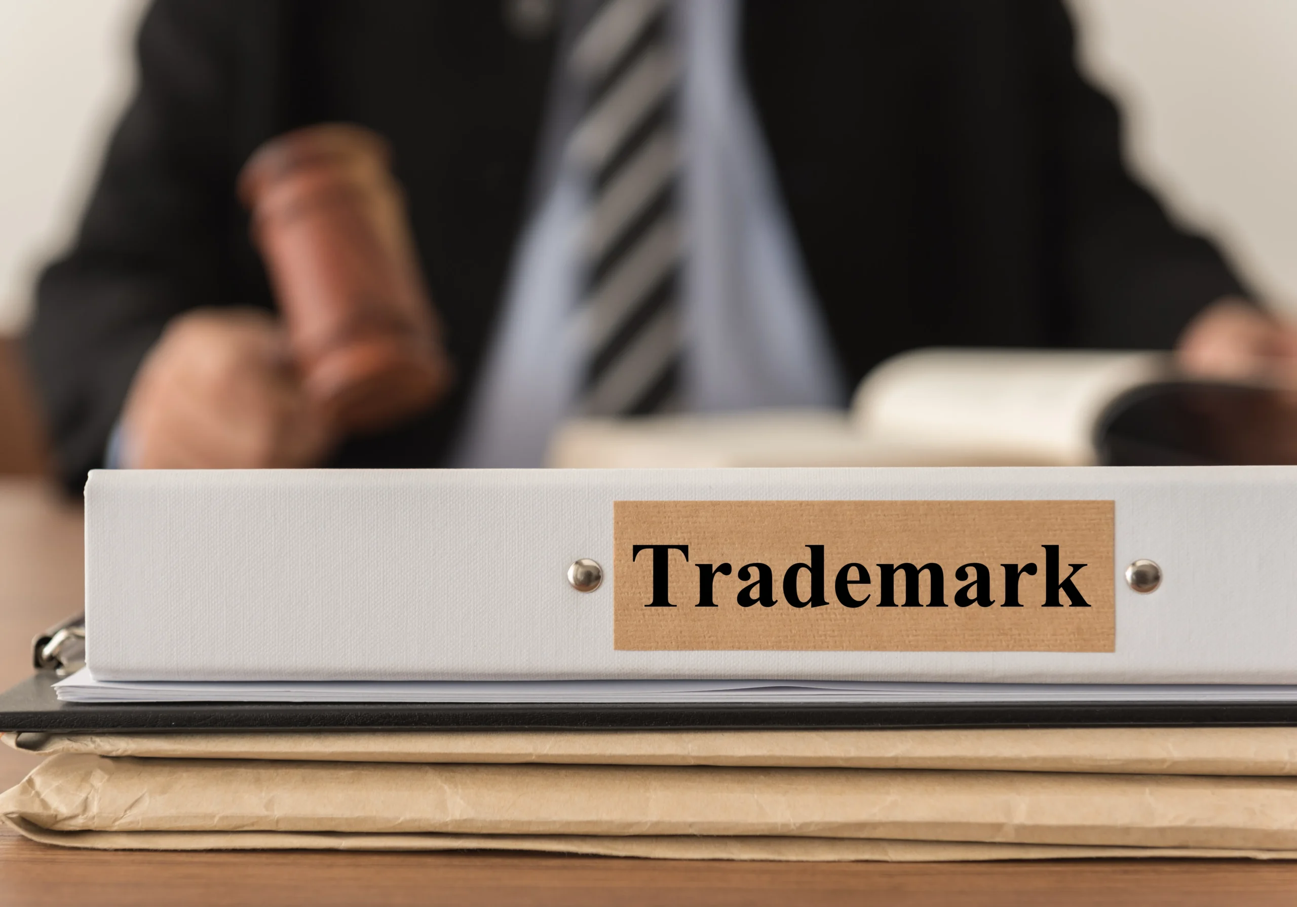 Close-up of a white binder labeled "Trademark" with a blurred person in suit holding a gavel in the background.