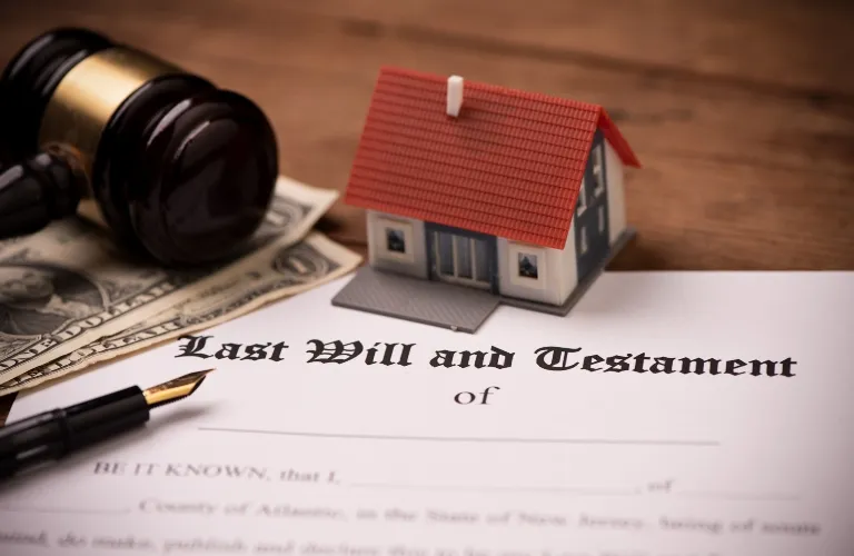 Document titled "Last Will and Testament" with a miniature house, gavel, pen, and US dollar bills on a wooden surface.