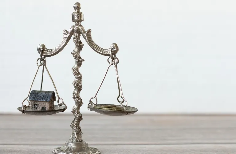 Silver balance scale with a miniature house on one side and coins on the other side.