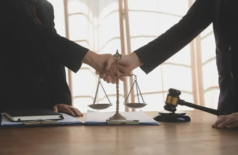 Two individuals in dark suits shaking hands over a wooden desk with a balance scale and judge's gavel.