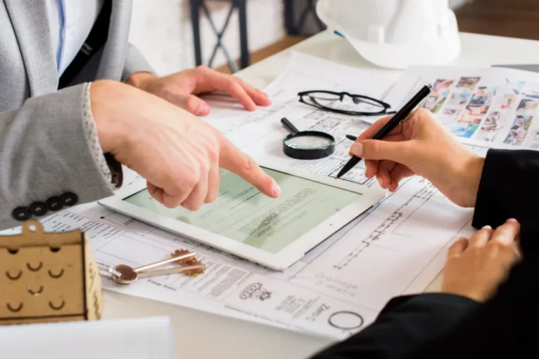 Two professionals reviewing documents on a tablet over architectural blueprints with keys, magnifying glass, and a white hard hat.