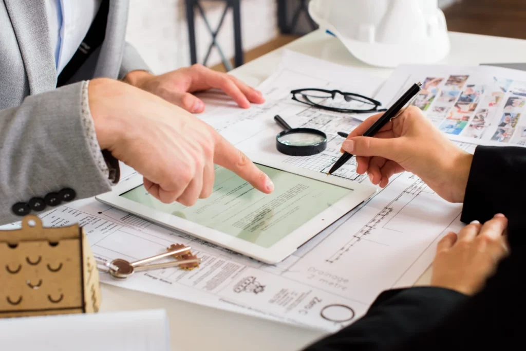 Two professionals reviewing documents on a tablet over architectural blueprints with keys, magnifying glass, and a white hard hat.